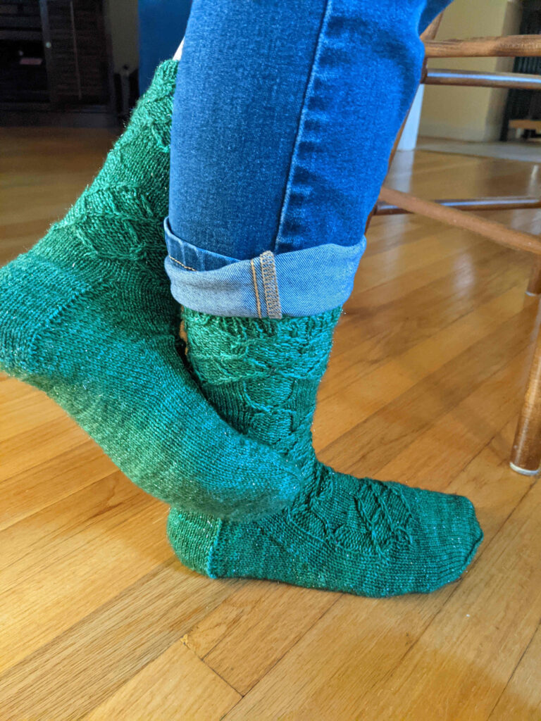 Pair of feet in Ebla Socks knit in Anzula Nebula yarn, color Fern. One toe curls around the other ankle.