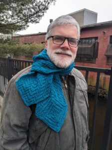 Man wearing Herringbone Hexagonal Scarf over a leather jacket while standing near the railing of a canal.