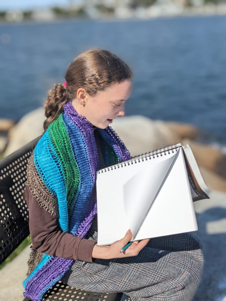 A young woman sits on a park bench in front of a bay with a large sketchbook, wearing a turtleneck and the Color Conduit shawl.
